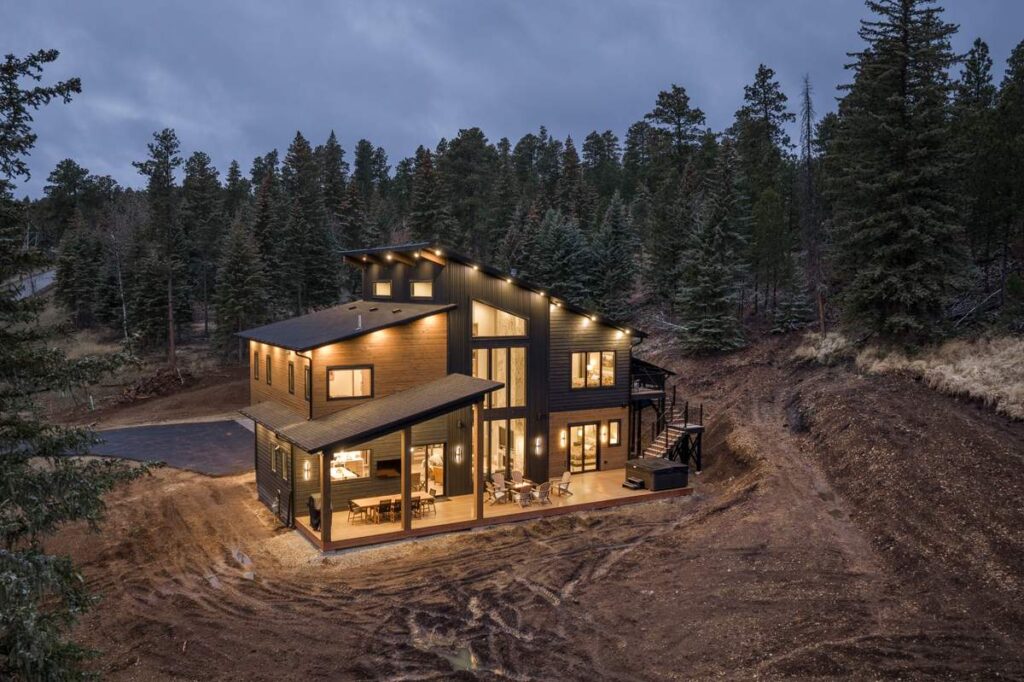 Modern mountain home exterior at dusk with warm lighting, surrounded by pine forest